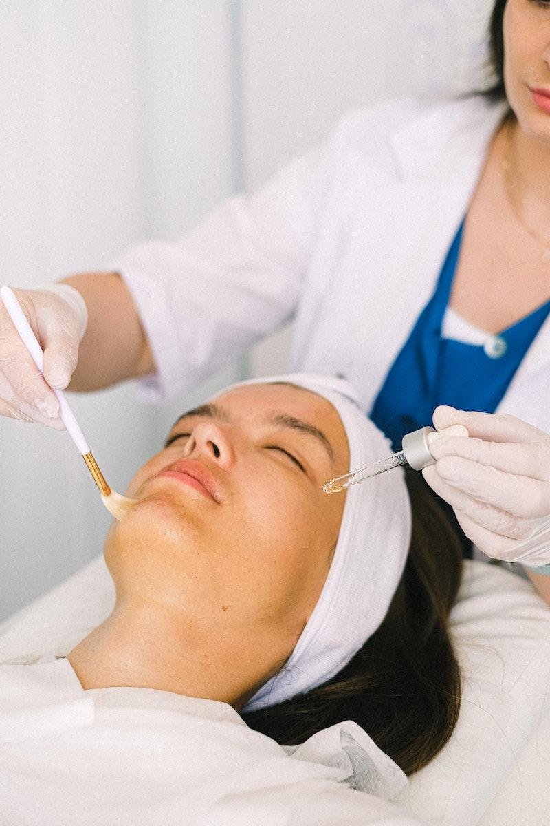 Young female cosmetologist in medical robe applying a phenol peel on female client's face skin lying with eyes closed in light modern spa salon