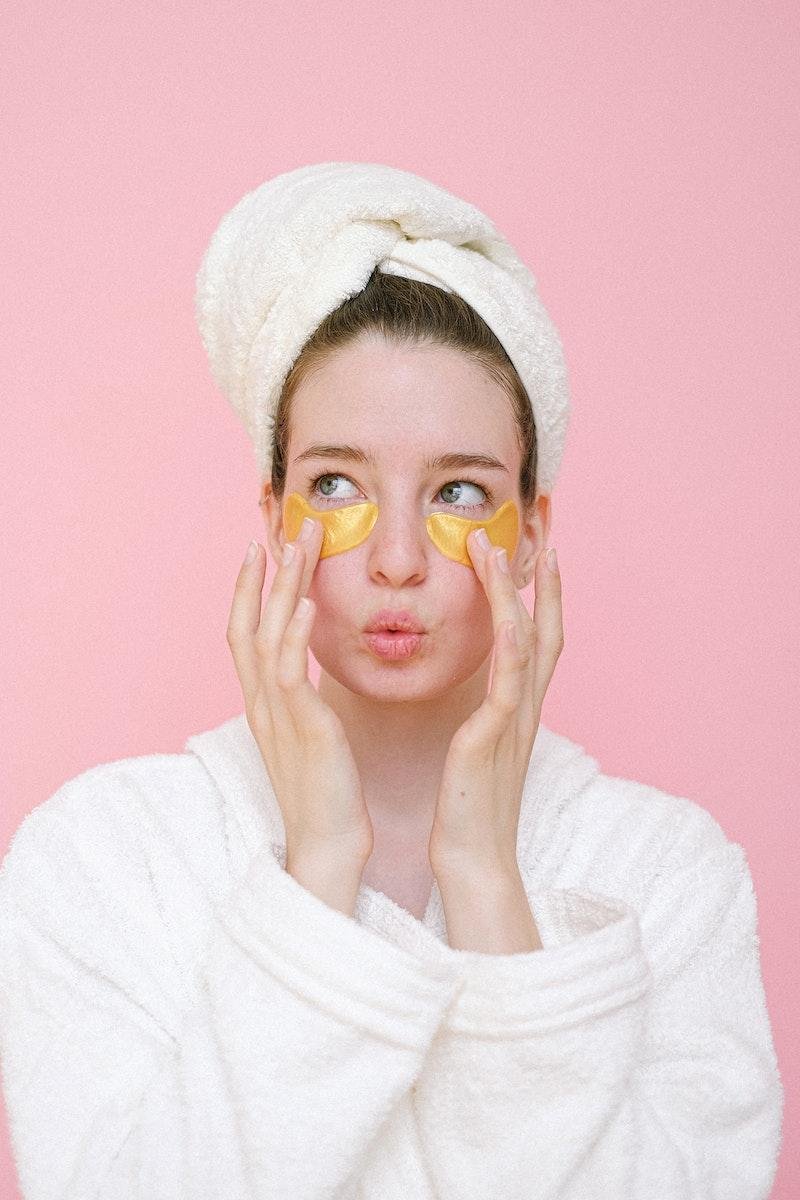 young woman wearing white bathrobe and towel on head applying eye patches against pink background